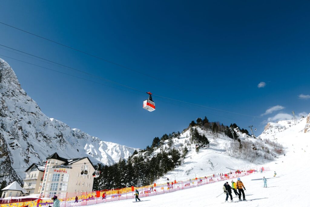 Red gondola cabin passing above skiers at the base of a snowy ski area under blue sky